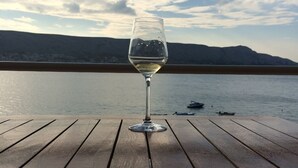 A glass of white wine on a wooden table with a view of a calm lake and mountains in the background. - Hotel Belveder (Pag)