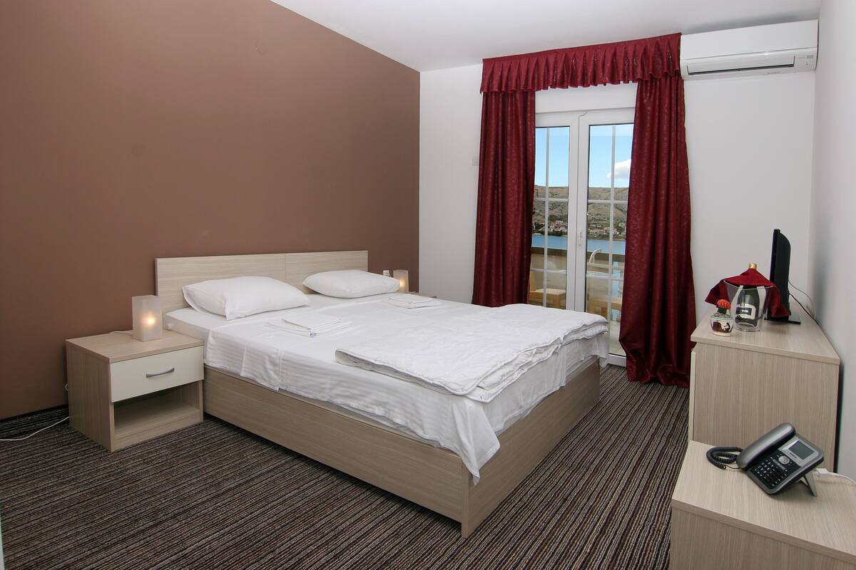 A hotel room with a bed, a nightstand, a desk with a phone, and a window with curtains.
