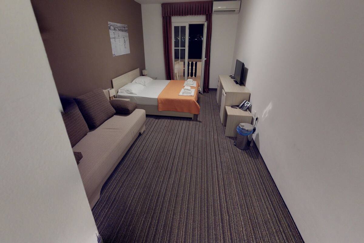 A hotel room with a bed, a sofa, a desk with a phone, and a trash bin.