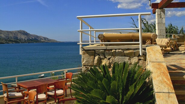 A terrace with wooden furniture, a stone wall, and a view of the sea. - Hotel Belveder (Pag)