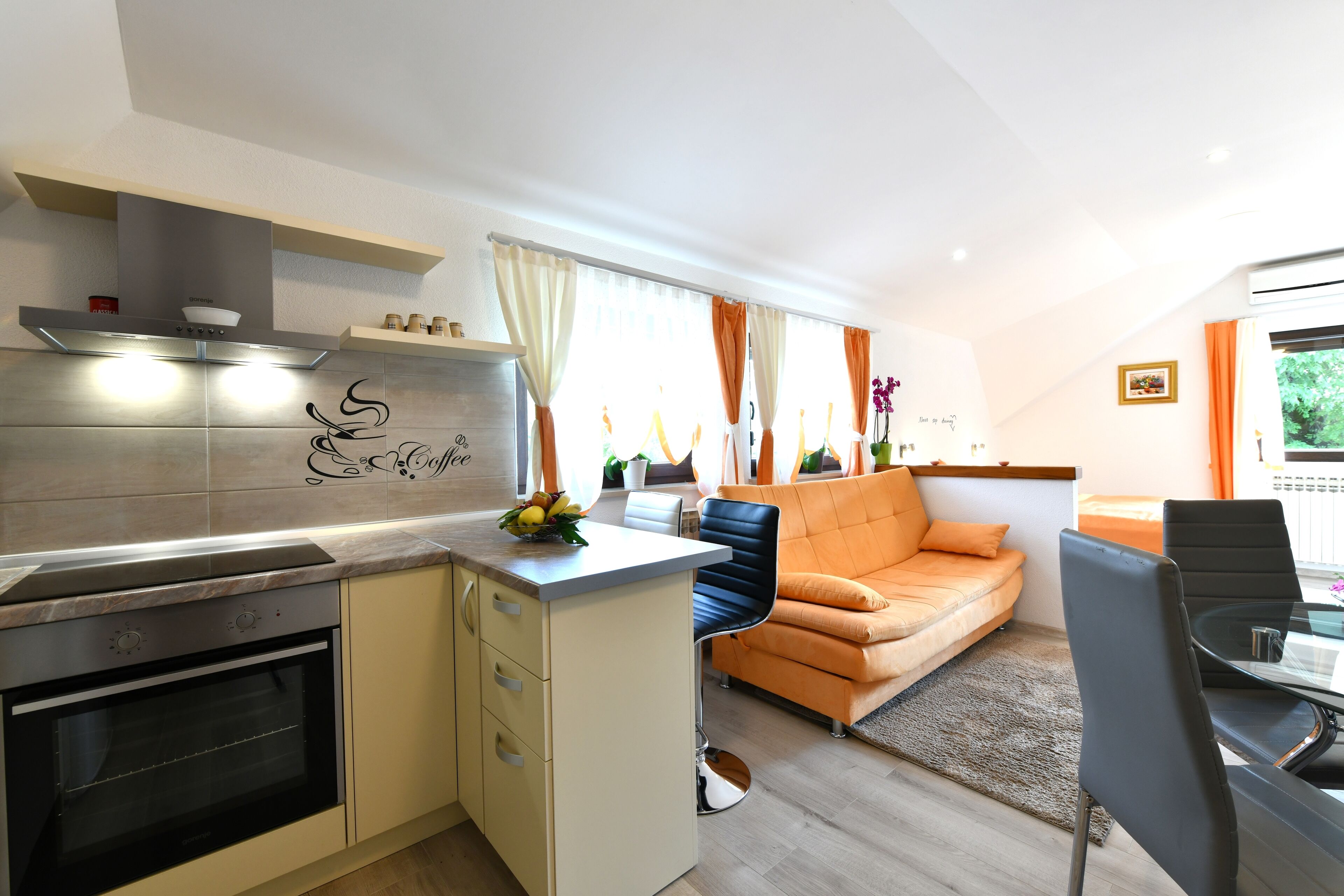 A modern kitchen with a built-in oven, a dining area with a glass table, and an orange sofa.