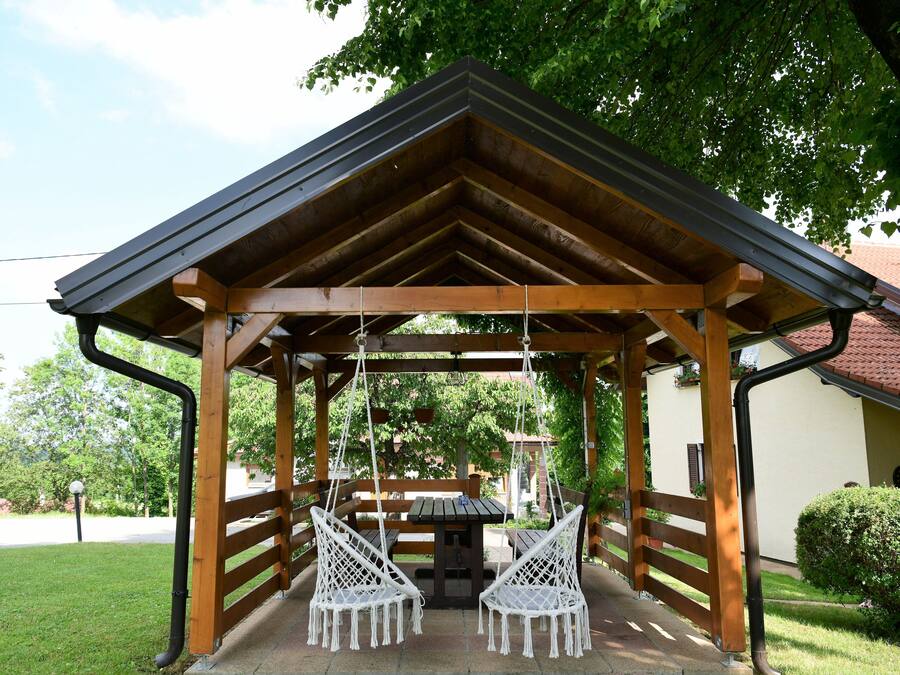 A wooden gazebo with two white swing chairs and a picnic table.