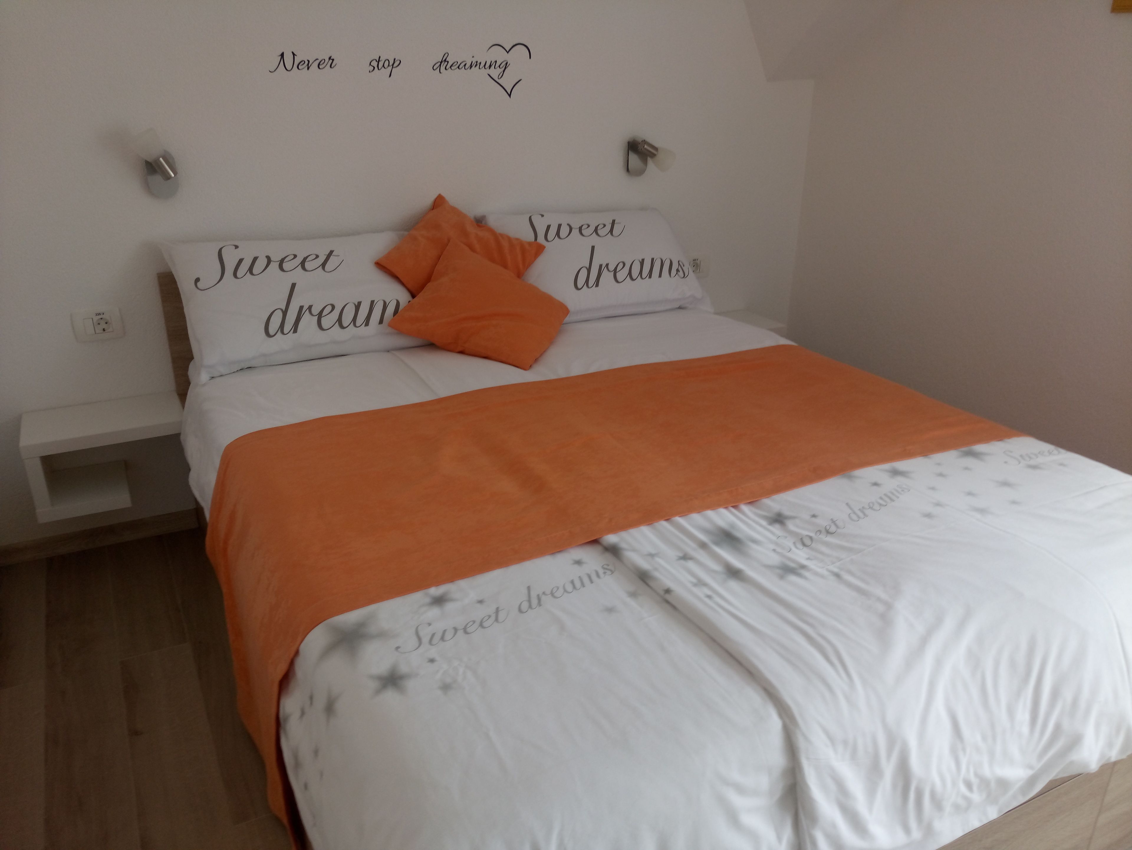 A neatly made bed with a white and orange bedspread, two pillows, and a wall with the text "Sweet dreams" above it.