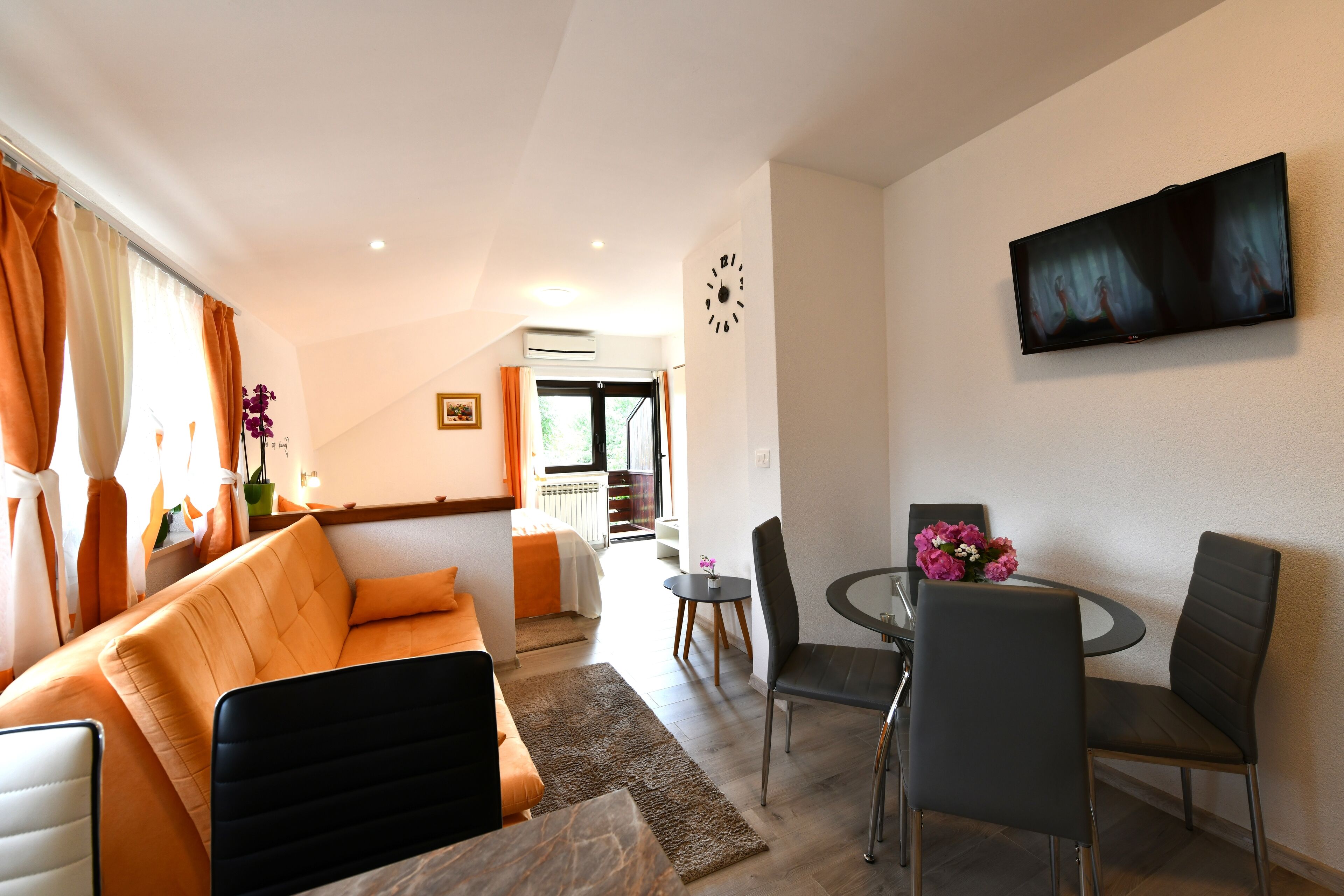 A modern living room with a sofa, a dining table with chairs, and a flat-screen TV mounted on the wall.