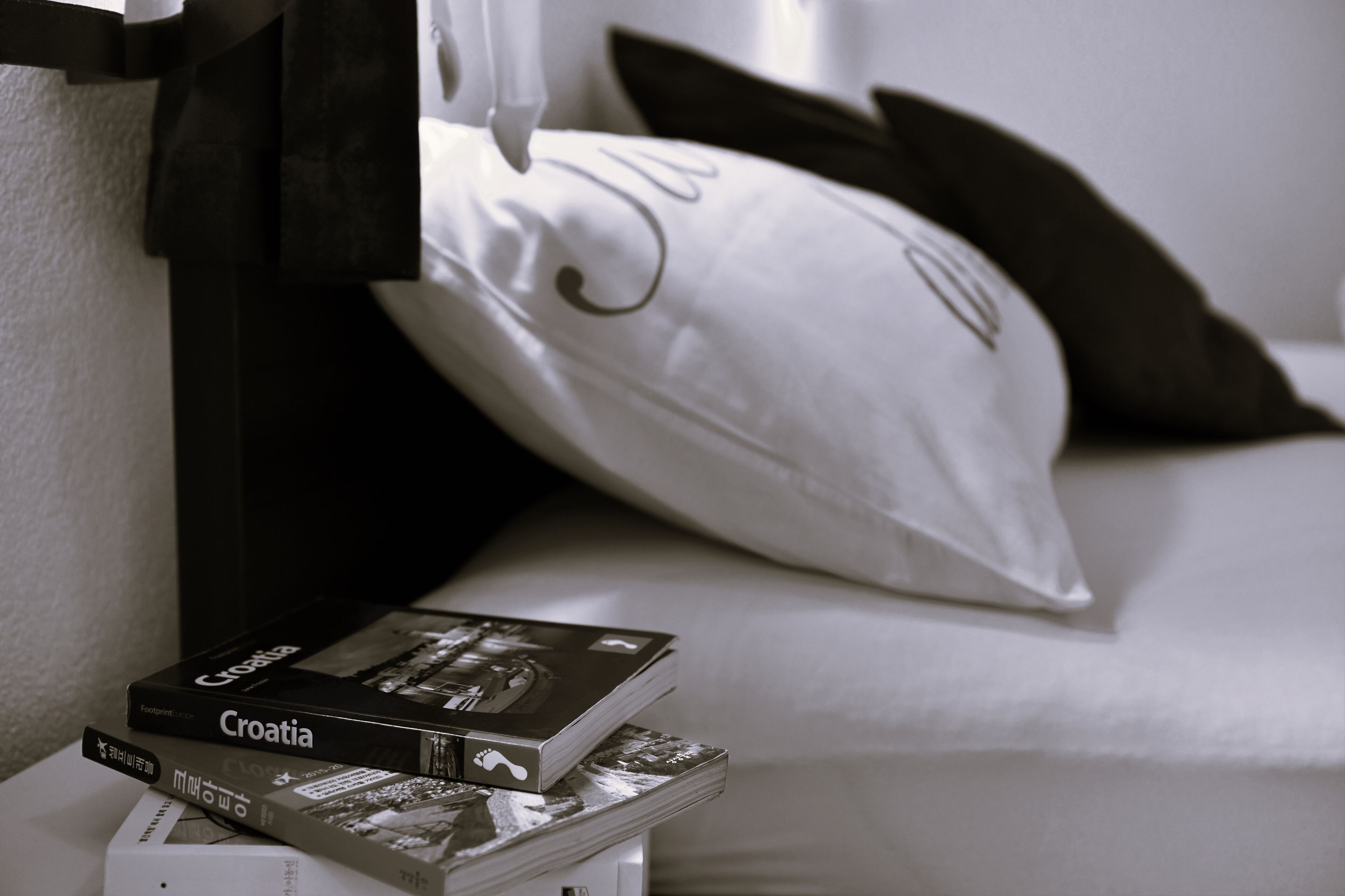A neatly arranged stack of books on a bedside table, a pillow with a decorative design, and a dark-colored headboard.