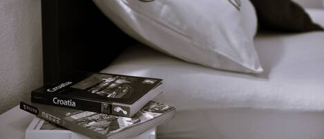 A neatly arranged stack of books on a bedside table, a pillow with a decorative design, and a dark-colored headboard.