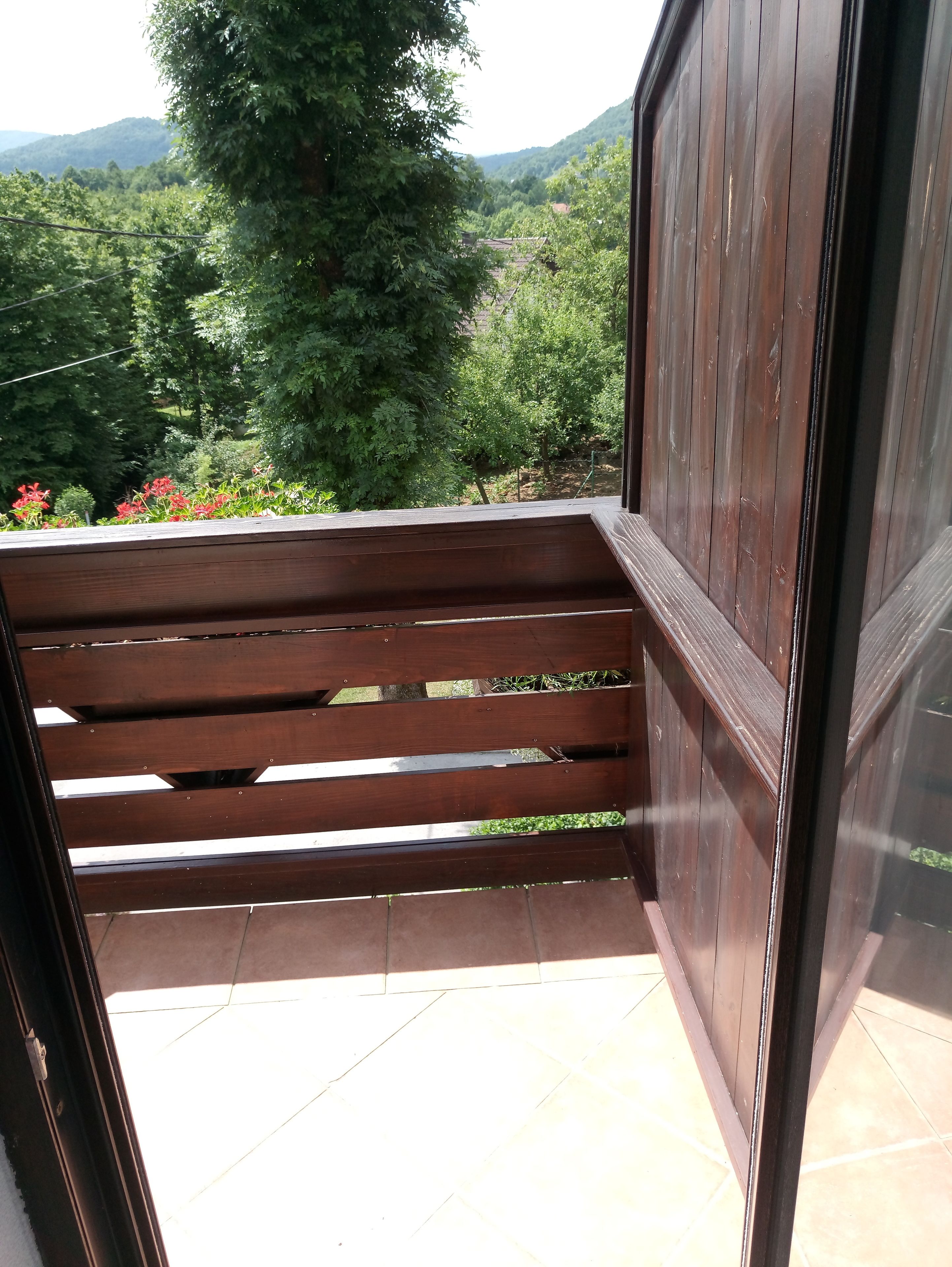 A balcony with a wooden bench, a glass door, and a view of a green landscape with trees and hills.
