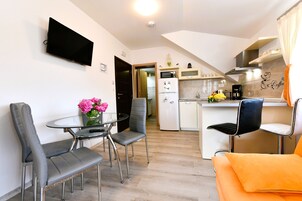 Una cocina moderna con una televisión de pantalla plana, una zona de comedor con una mesa y sillas de vidrio, y un rincón de estar acogedor con un cojín naranja.