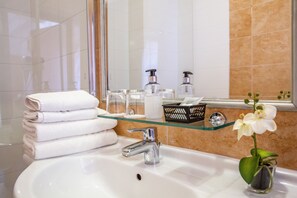 A bathroom with a white sink, a glass shelf with toiletries, a mirror, and a small orchid plant. - Fonte Hotel (Gyor)