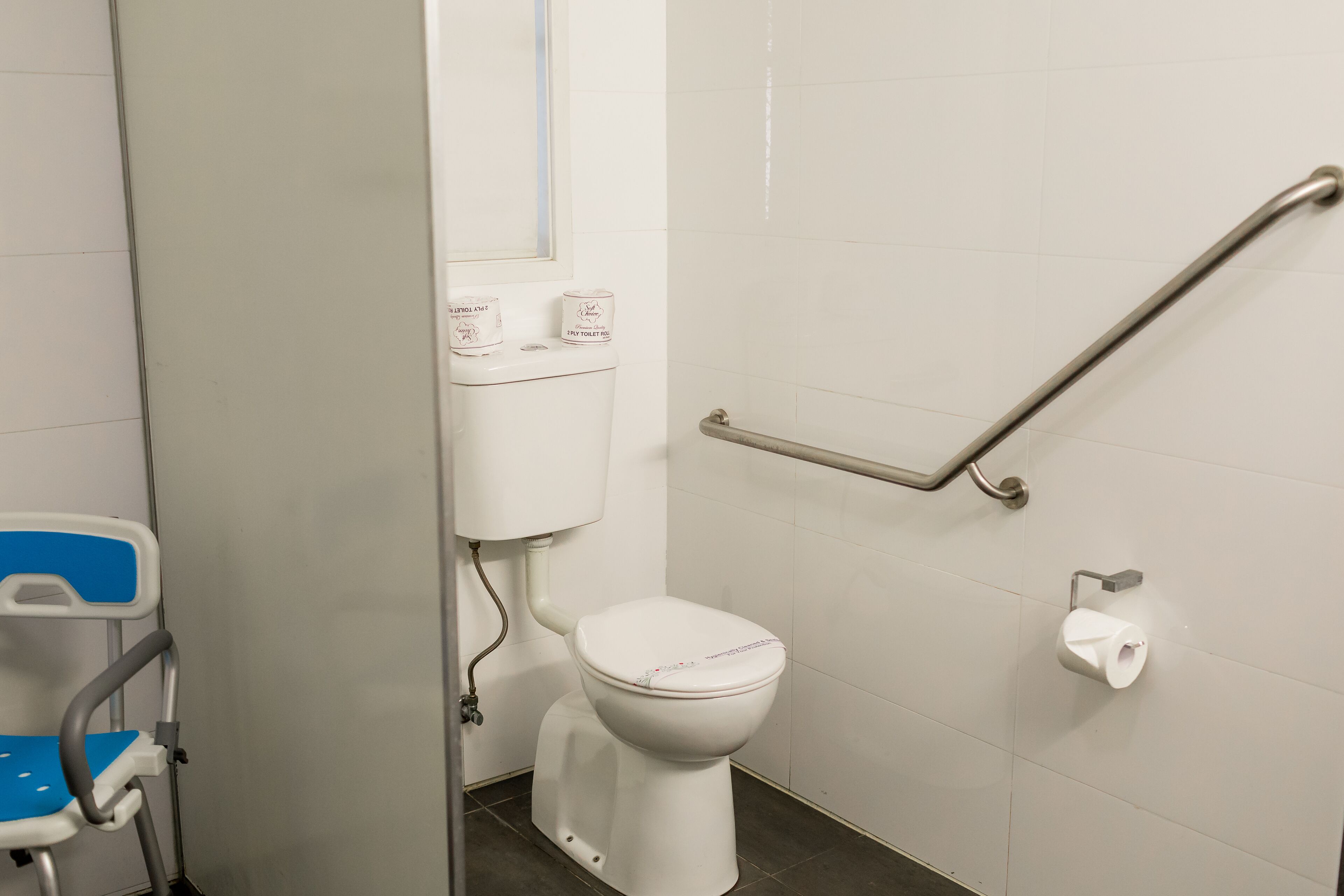 Un inodoro blanco con un asidero, una silla azul y dos rollos de papel higiénico en la pared.
