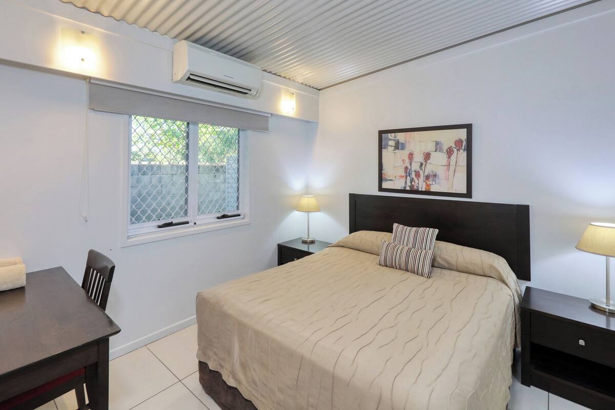 Une chambre à coucher comprenant un lit, des tables de chevet, un bureau et une fenêtre avec des stores.