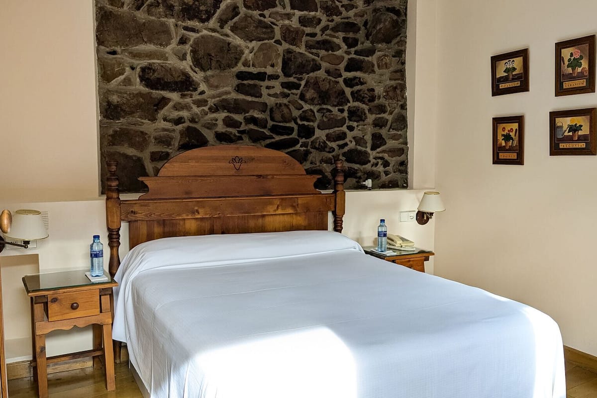 A bedroom with a stone wall, a wooden bed, two bedside tables with lamps, and four framed pictures on the wall.