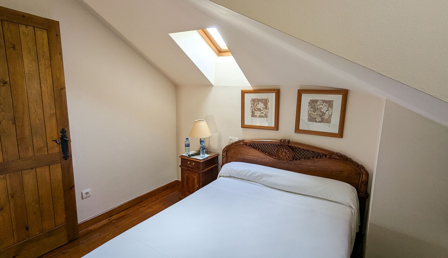 Een slaapkamer met een schuine plafond, houten deur, nachtkastje met een lamp, twee ingelijste foto’s en een bed met wit beddengoed.
