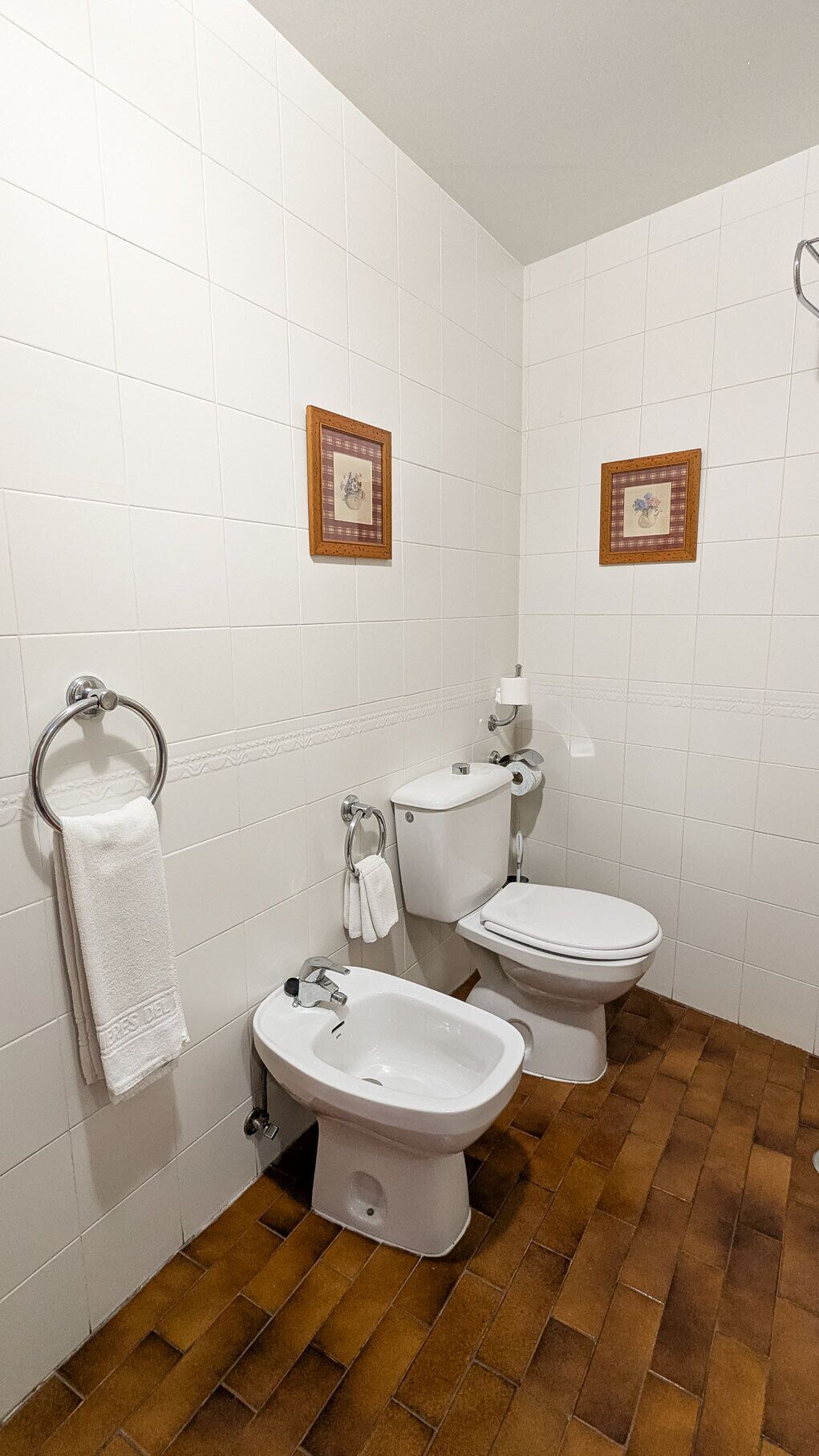 Un baño con paredes de azulejos blancos, dos inodoros, un bidé y un lavamanos. Hay dos cuadros en la pared.