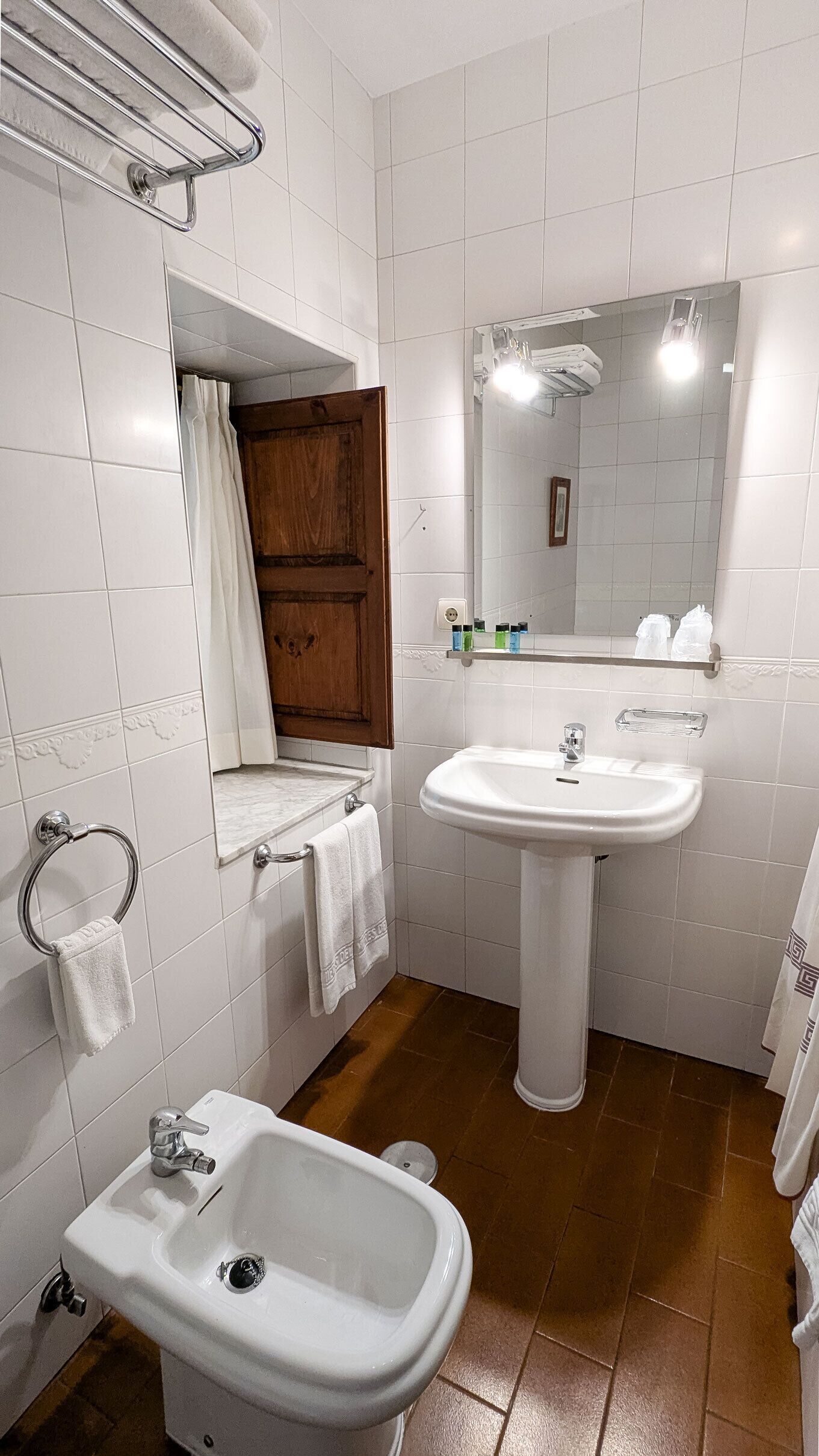 Een badkamer met witte tegels, een wastafel op voet, een bidet, een houten deur en een spiegel met verlichting.