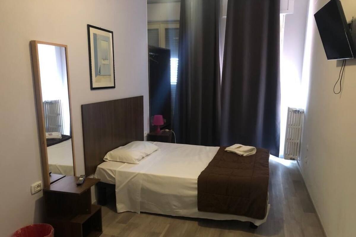 A hotel room with a single bed, a bedside table, a mirror, a TV mounted on the wall, and a window with curtains.