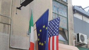 Entrada de un hotel con un letrero que indica el segundo piso y un piano. Se exhiben banderas de Italia, la UE y Estados Unidos.