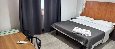 A hotel room with a bed, a wooden desk, a chair, and a remote control on the desk.