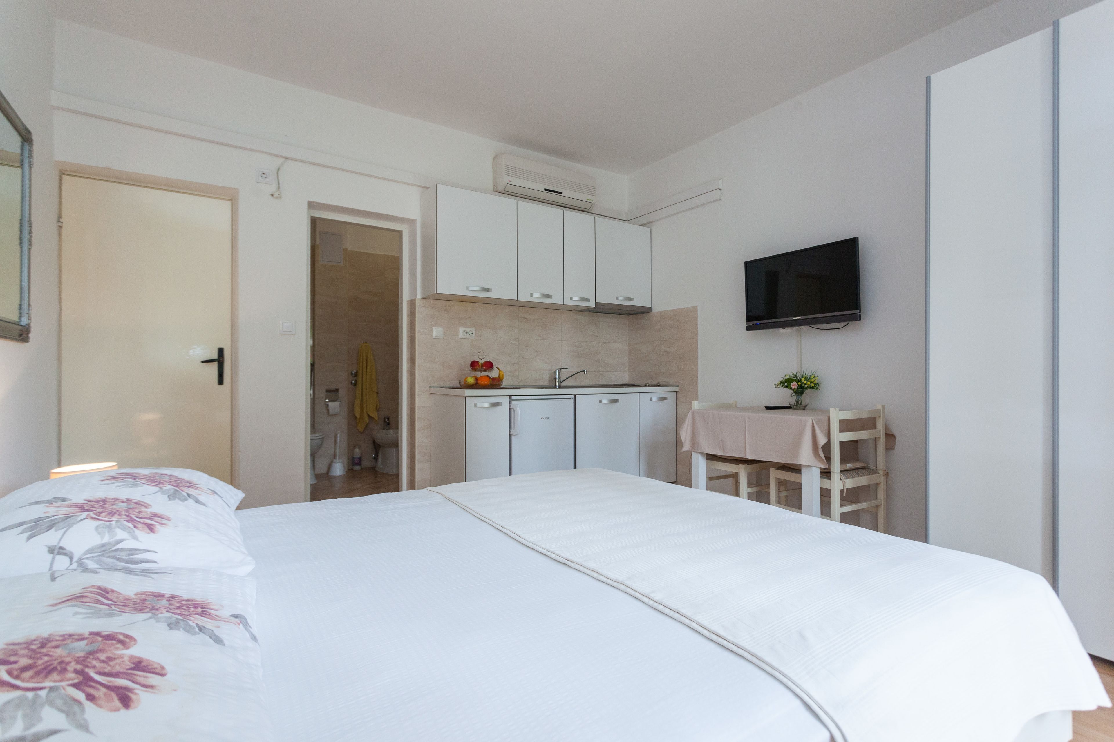 A bedroom with a bed, a small kitchenette, a TV mounted on the wall, and a bathroom visible through an open door.