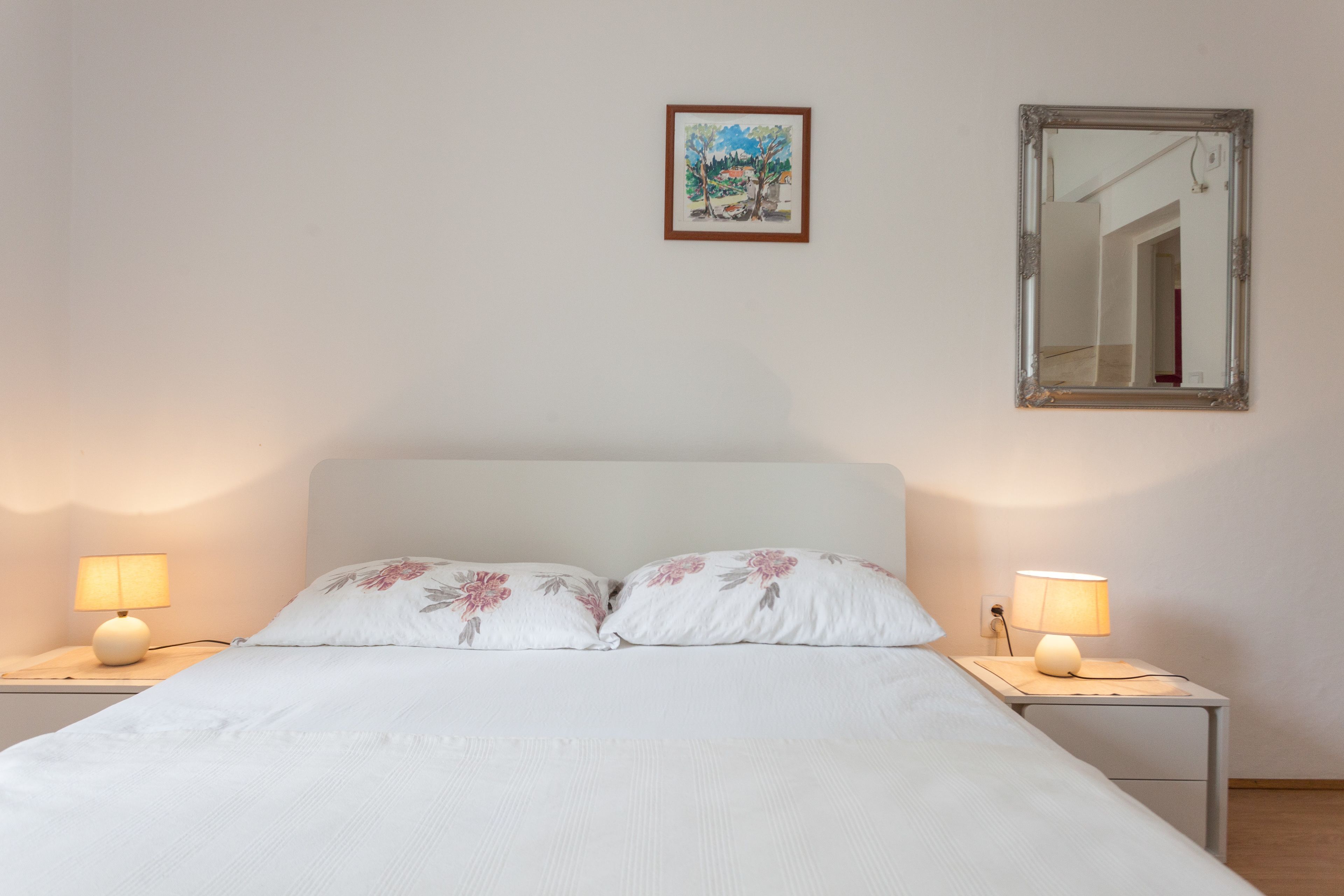 Een slaapkamer met een bed, twee nachtkastjes met lampen, een lijst met een schilderij en een spiegel aan de muur.
