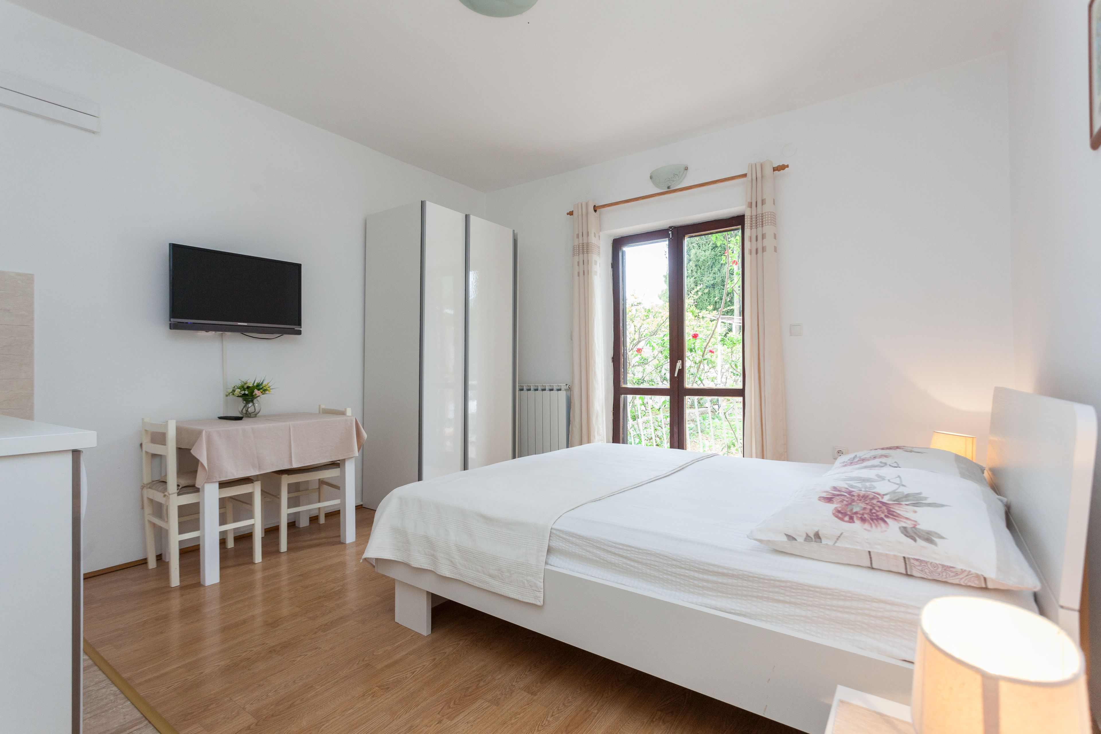 Un dormitorio con una cama, un televisor montado en la pared, un escritorio con una silla y una ventana con cortinas.