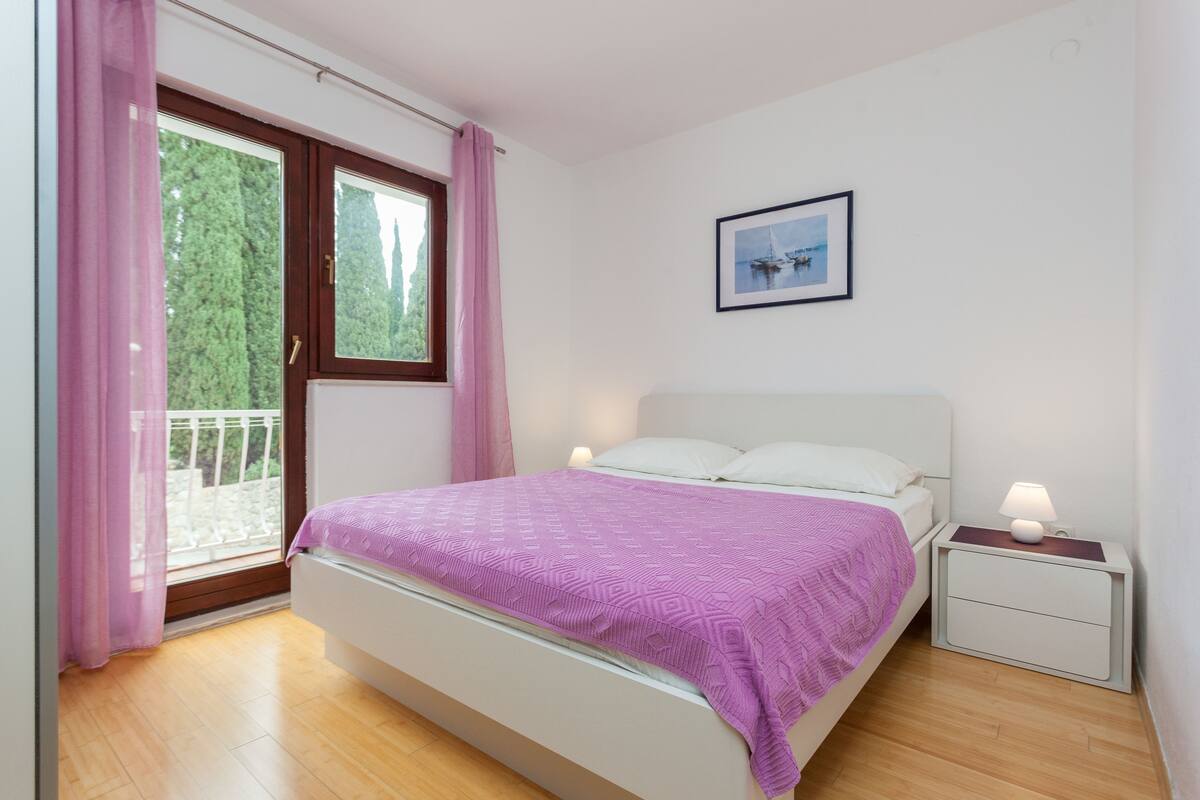 A bedroom with a bed, a nightstand with a lamp, a window with curtains, and a framed picture on the wall.