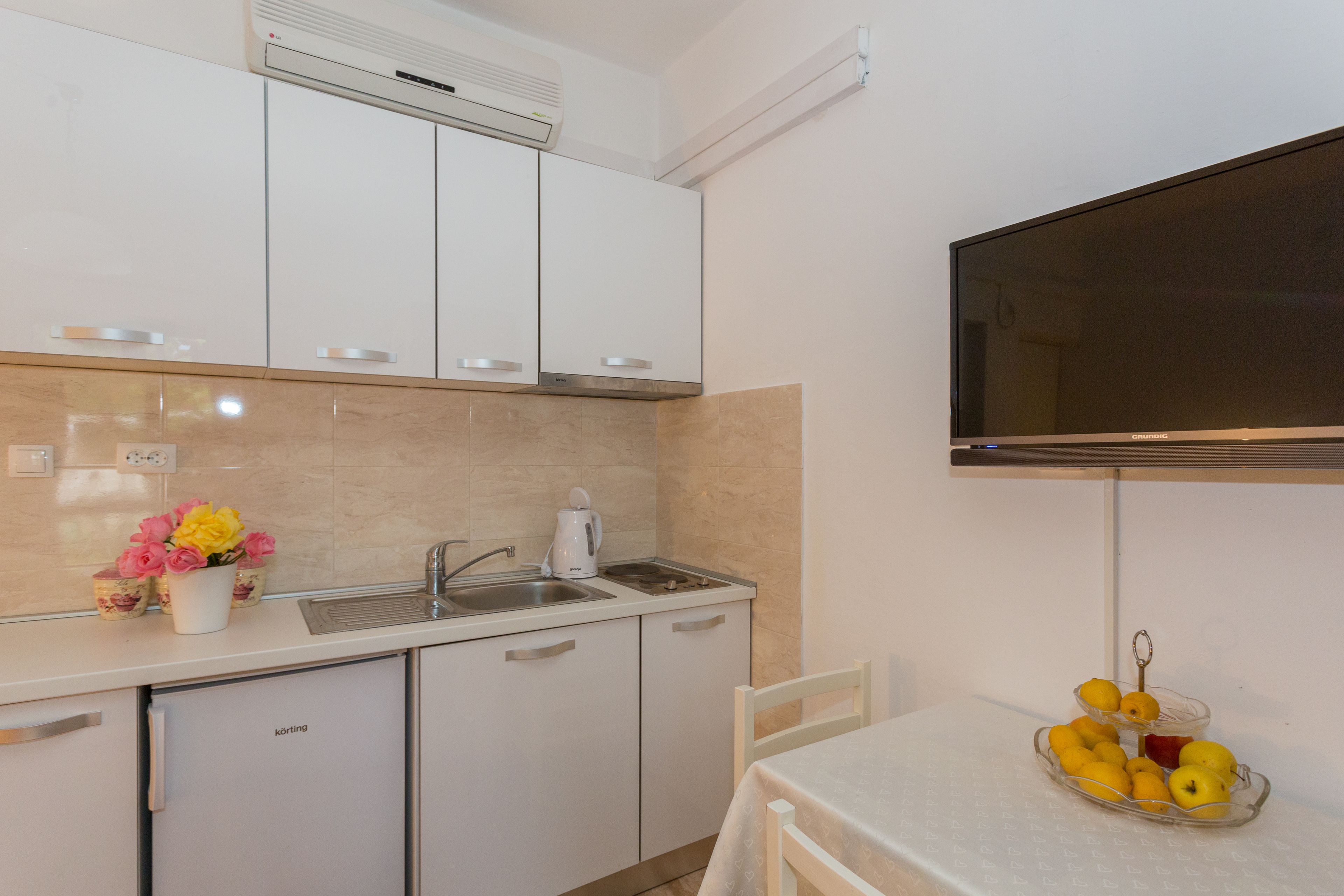 A compact kitchen with white cabinets, a sink, and a small refrigerator. There is a white table with a bowl of fruit and a vase of flowers on the counter.