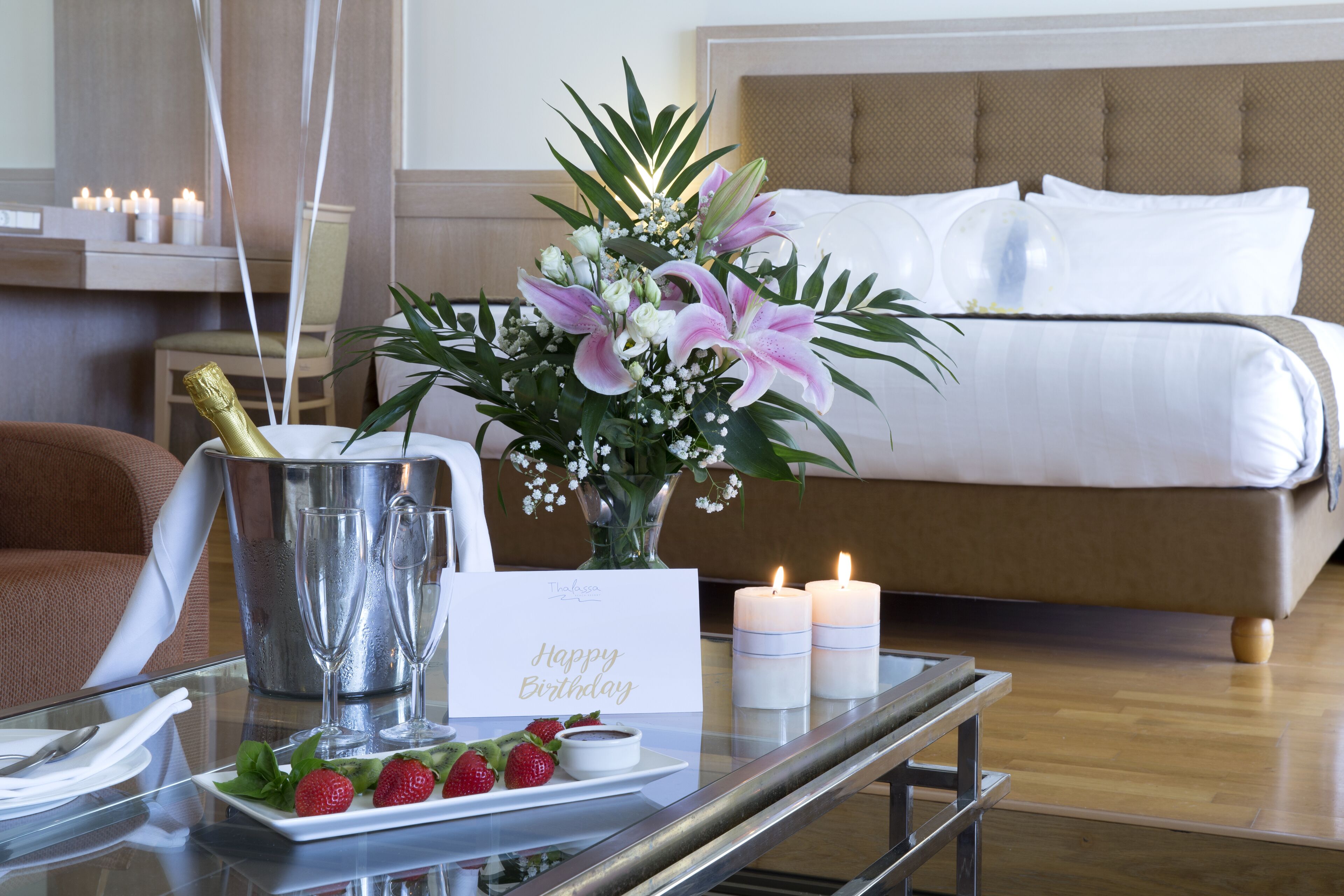 Ein Hotelzimmer mit Bett, Nachttisch mit Kerze, Blumenvase, einer Champagnerflasche und einer Geburtstagskarte.