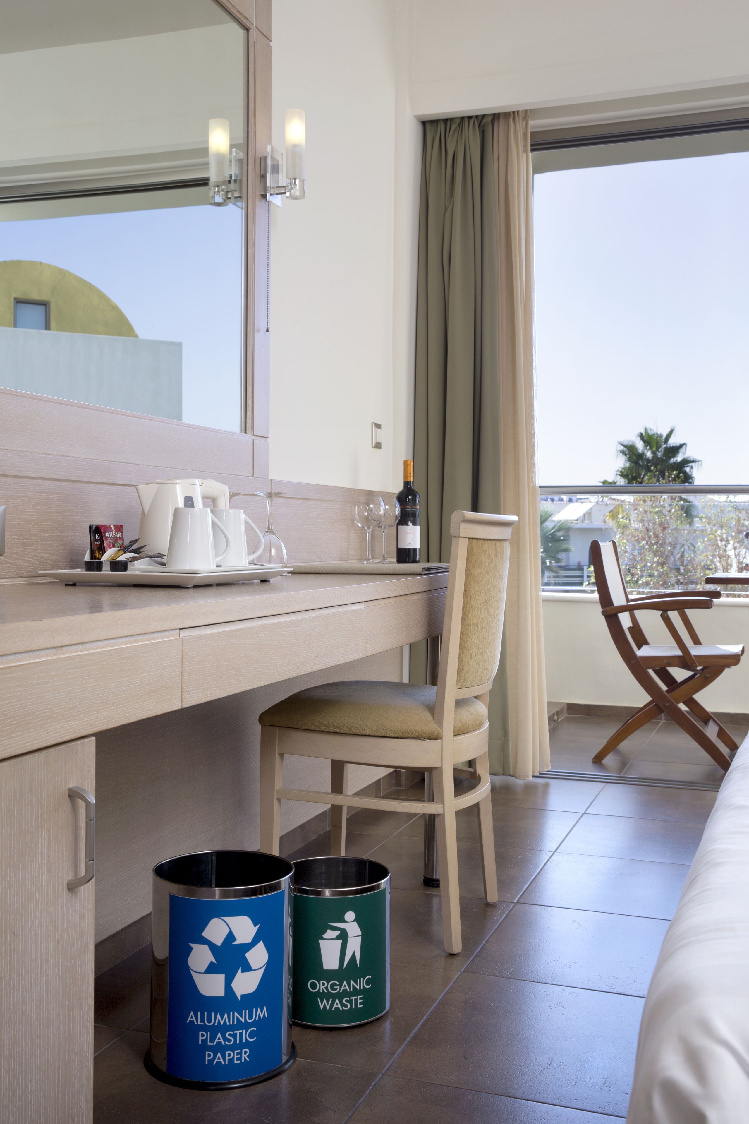 A hotel room with a wooden desk, a chair, a bed, and two waste bins labeled for recycling.