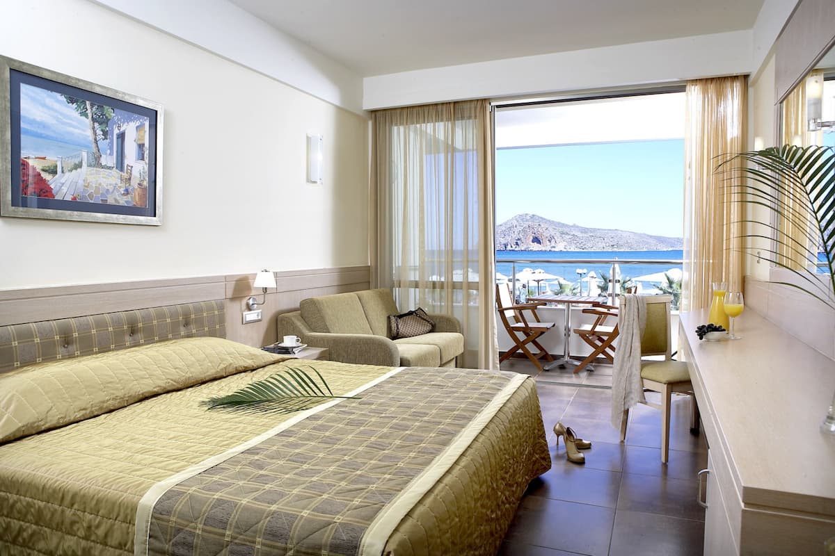 Quarto de hotel com cama, sofá, mesa de jantar e cadeiras, e vista para o mar.