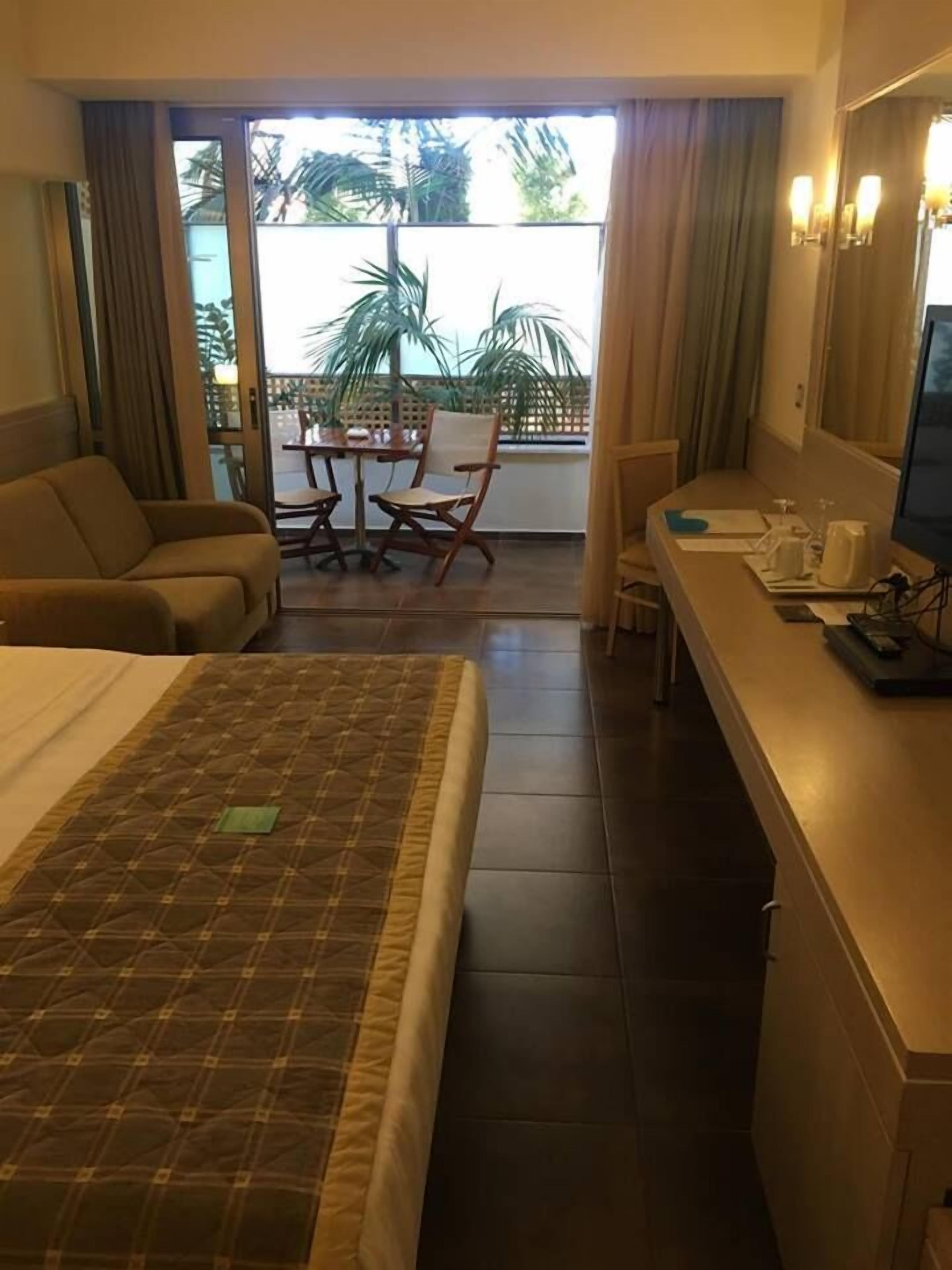 A hotel room with a bed, a sofa, a dining table with chairs, a small kitchen area, and a sliding glass door leading to an outdoor area with plants.