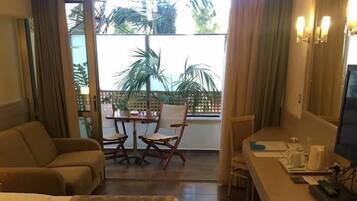 A hotel room with a bed, a sofa, a dining table with chairs, a small kitchen area, and a sliding glass door leading to an outdoor area with plants.