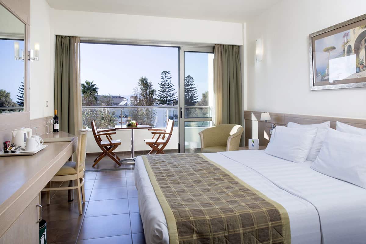 Quarto de hotel com uma cama grande, uma mesa com cadeira, varanda com mesa e cadeiras e um quadro na parede.