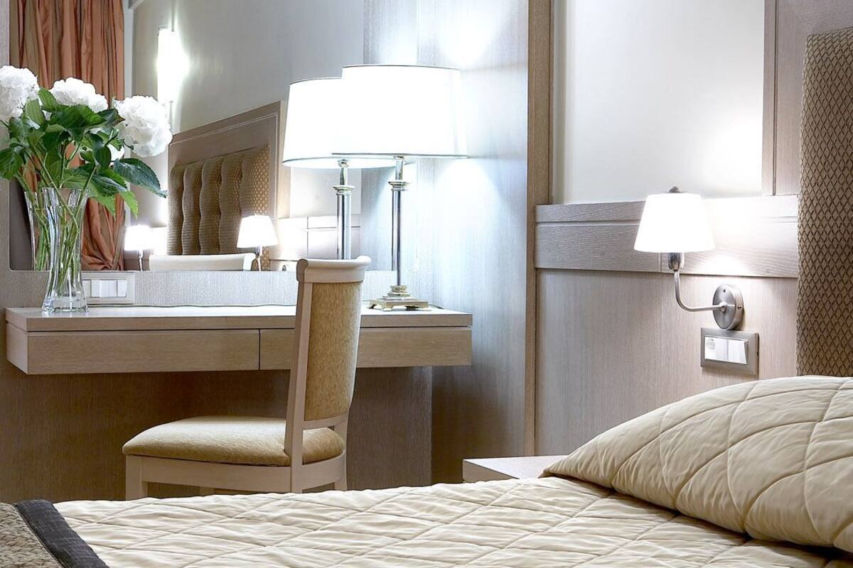 Quarto de hotel com cama, escrivaninha, cadeira, abajur e um vaso com flores.