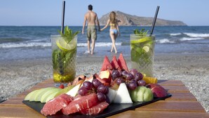 Een tafel met een fruitschaal en twee drankjes op een strand, met een stel in de achtergrond.