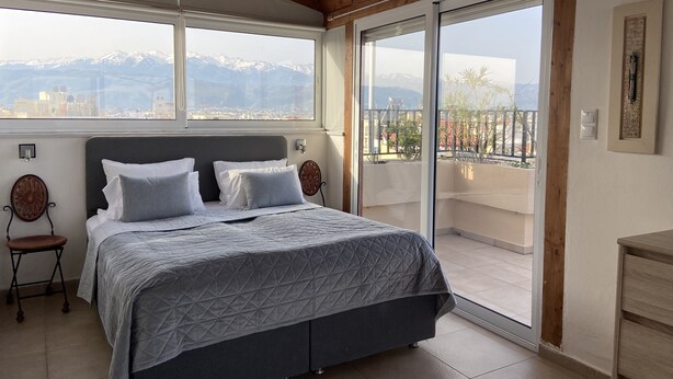 Une chambre avec un lit, un plafond en bois, une fenêtre donnant sur la ville et une porte-fenêtre coulissante qui mène à un balcon.