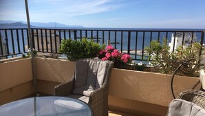 Un balcon agrémenté de meubles en osier, d’une table en verre et offrant une vue sur la mer et les montagnes.