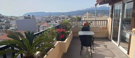 A balcony with a table and chairs, a potted plant, and a view of a cityscape with mountains in the distance.