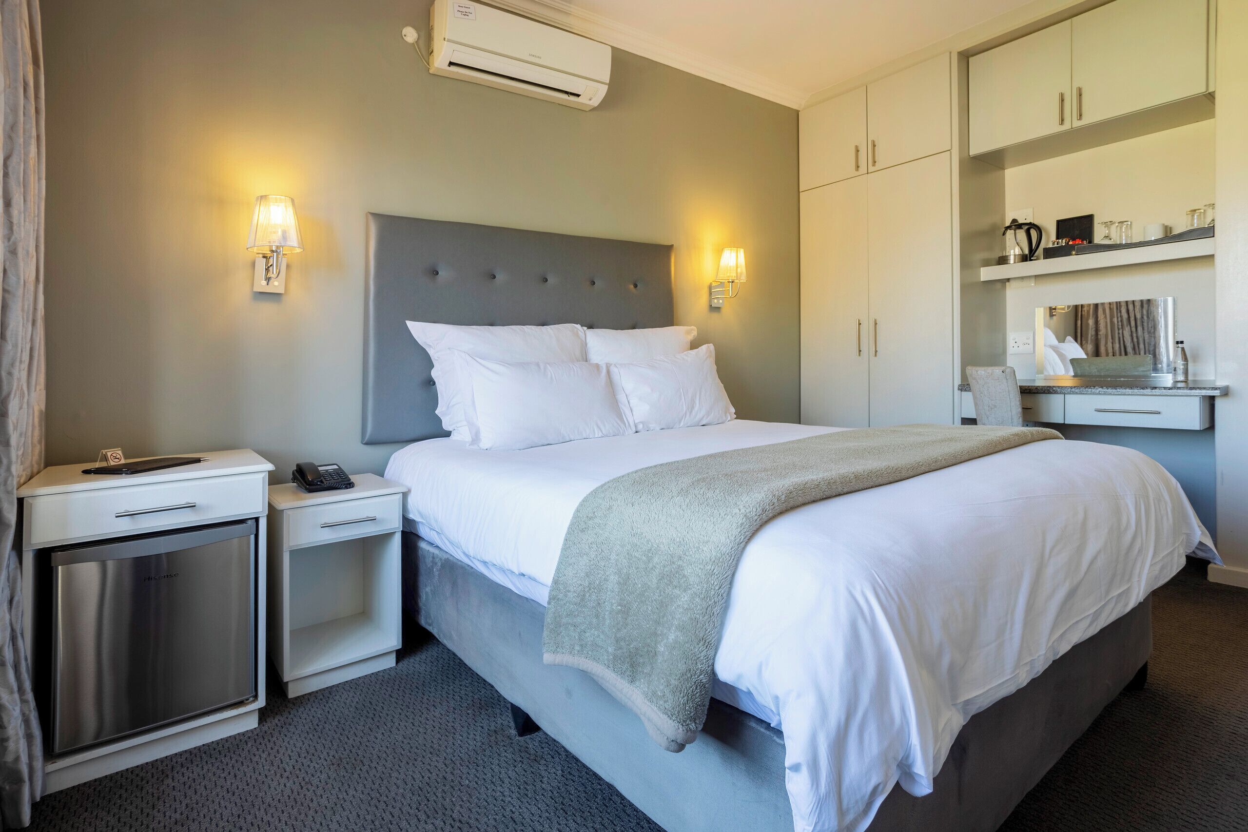 A hotel room with a bed, bedside tables, a small cabinet, and a wall-mounted shelf with items.
