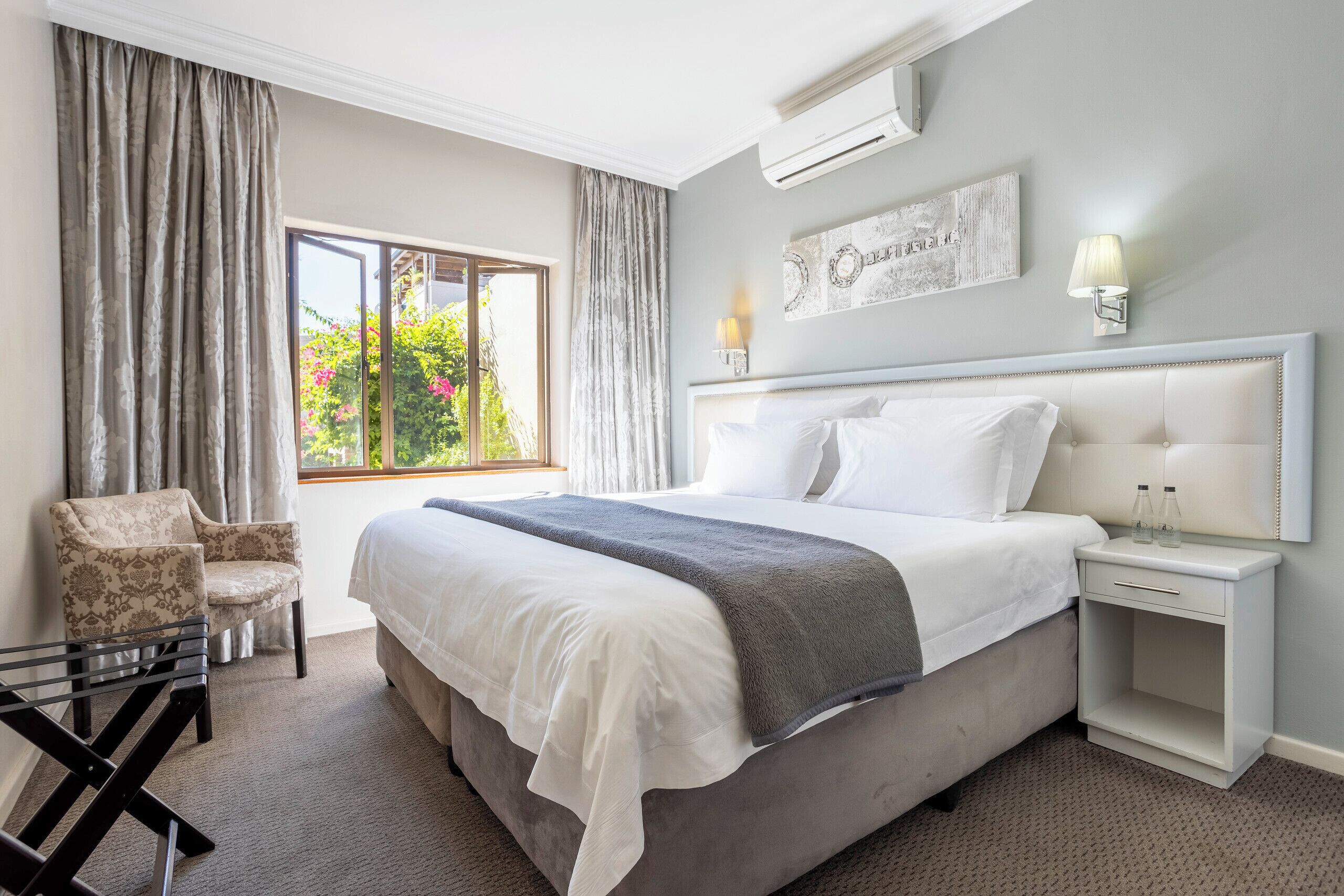 Una habitación de hotel ordenada con una cama grande, una silla, una mesita de noche y un ventanal con cortinas.
