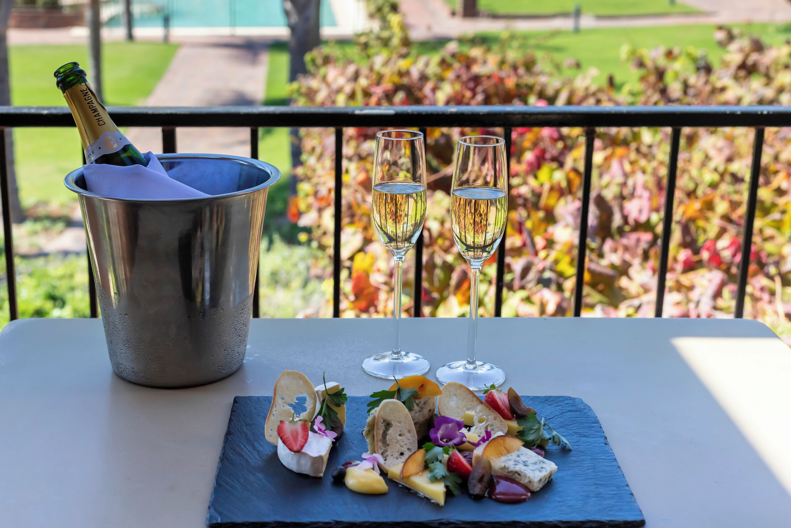 Een tafel met een fles champagne in een metalen emmer, twee champagneglazen en een schoolbord met verschillende kazen, fruit en bloemen.