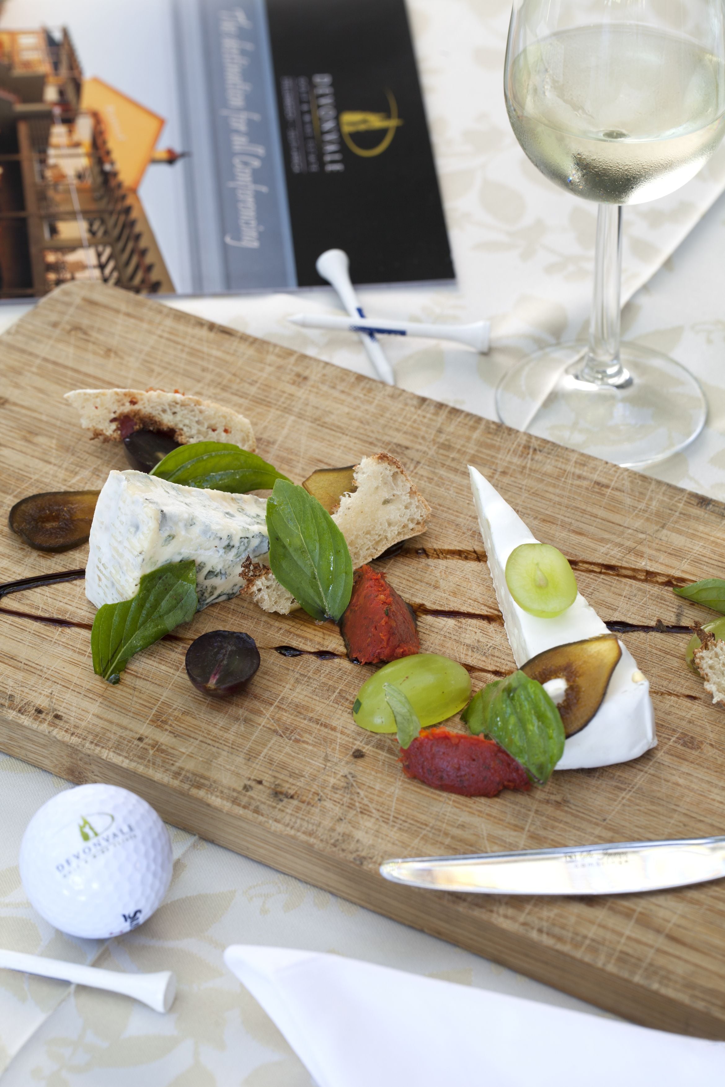 A wooden board with various cheeses, grapes, and bread, accompanied by a wine glass and a golf ball.