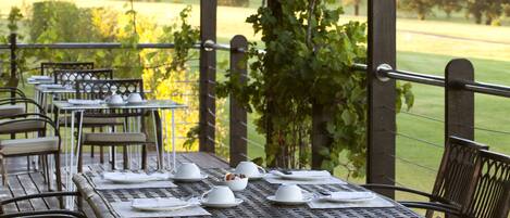 Un área de comedor al aire libre techada con mesas y sillas, una pérgola cubierta de enredaderas y vistas a un campo cubierto de hierba.