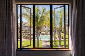 A window with a view of a well-maintained garden, including trees and a swimming pool.