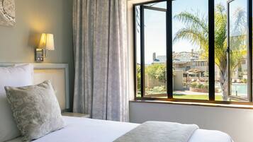 Quarto de hotel com uma cama, abajur, janela com vista para uma piscina e edifícios.