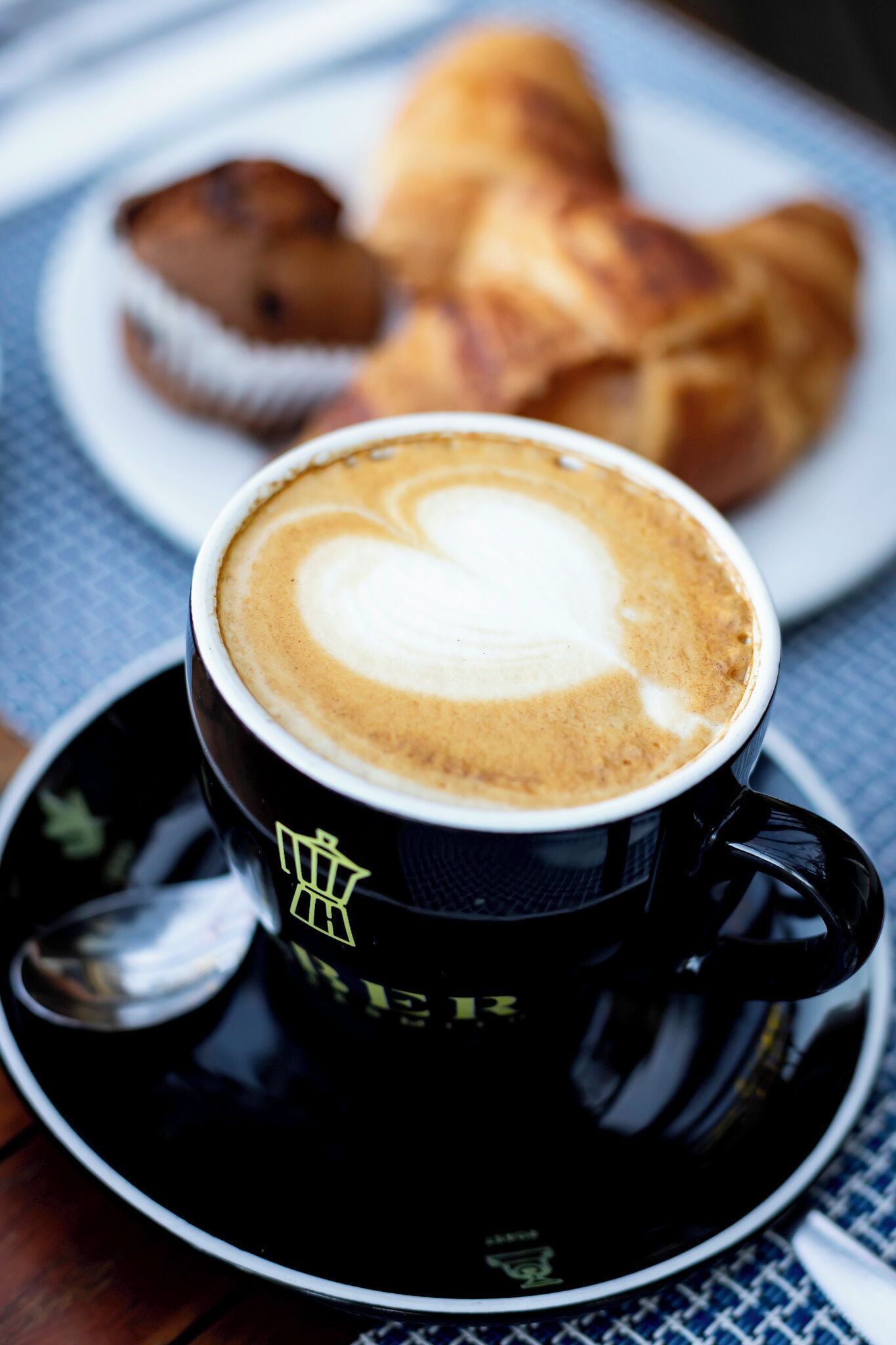 A cup of coffee with a heart-shaped latte art on a black cup with a logo, placed on a saucer. In the background, there is a plate with croissants.