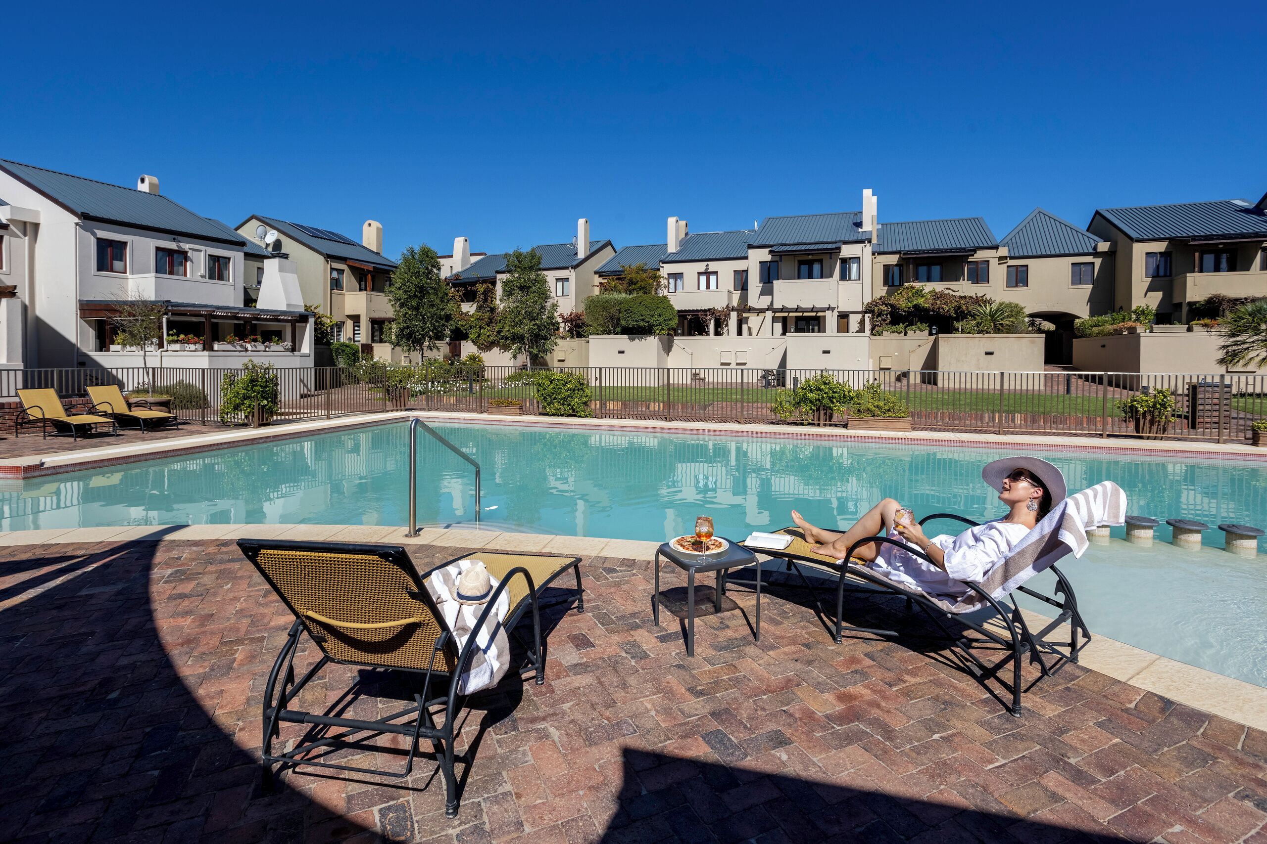 Un área junto a la piscina con sillones de descanso, una mesita con una bebida y vistas a edificios residenciales al fondo.
