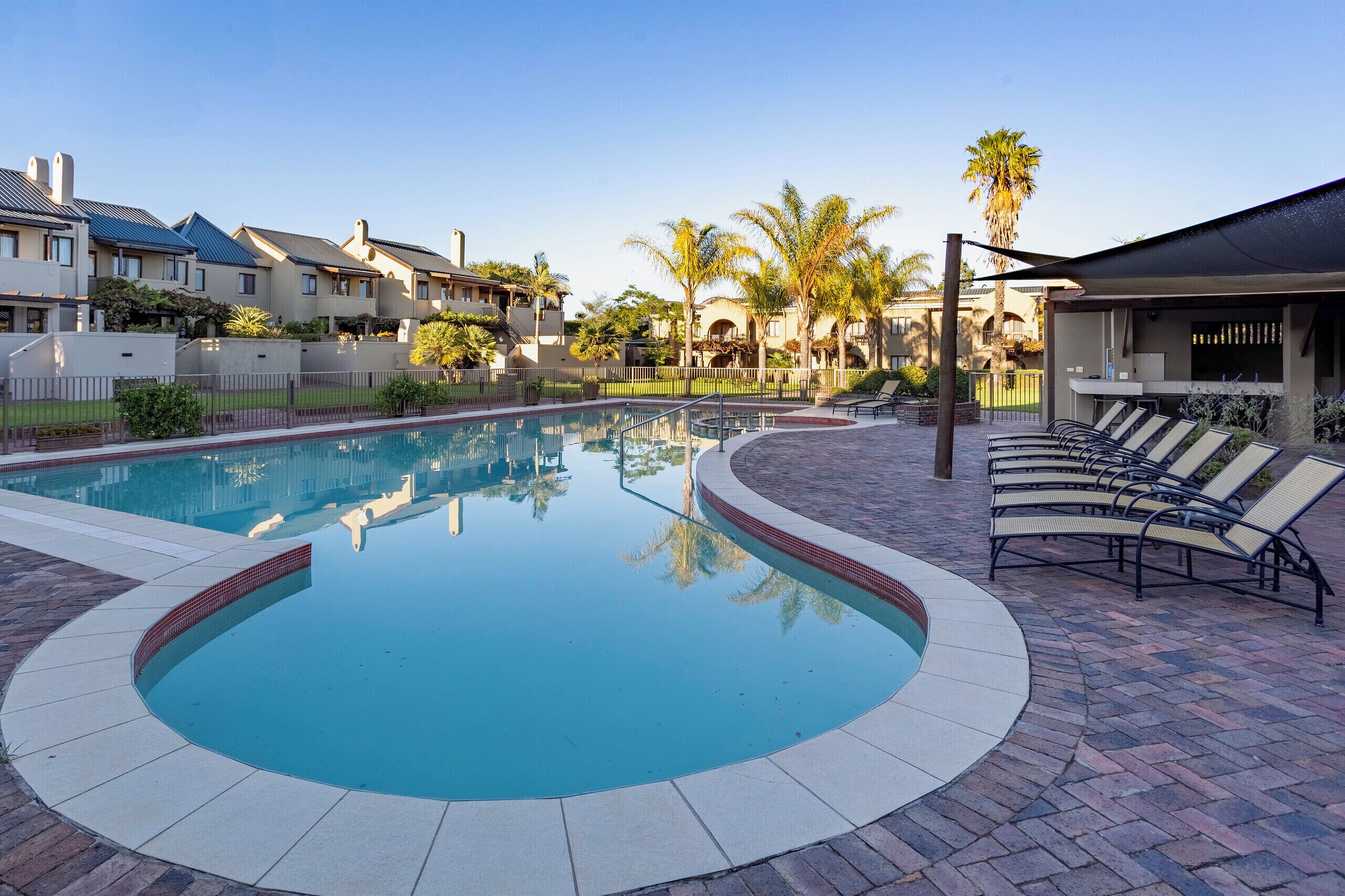 A swimming pool with a curved edge, surrounded by a brick patio. There are lounge chairs and a covered area with a table and chairs.