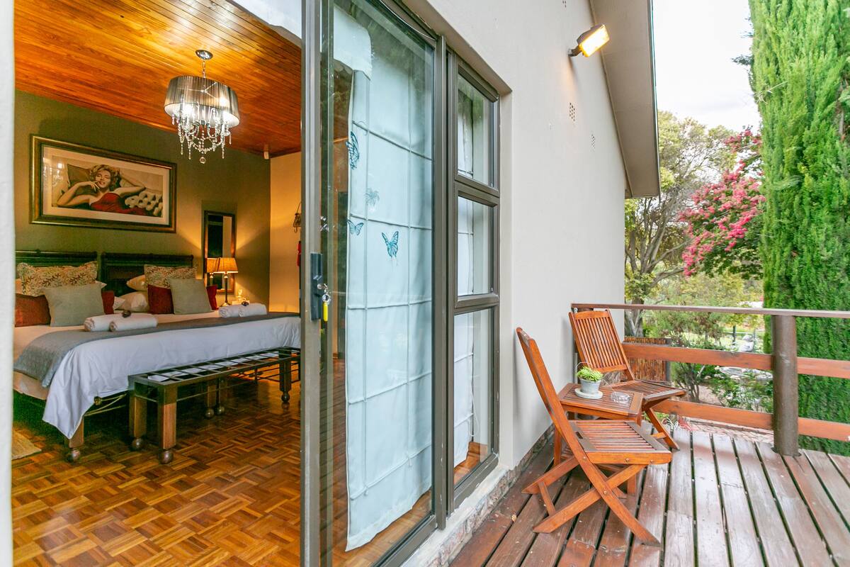 A bedroom with a bed, bedside table, and a chandelier. There is a balcony with a wooden railing and a chair.
