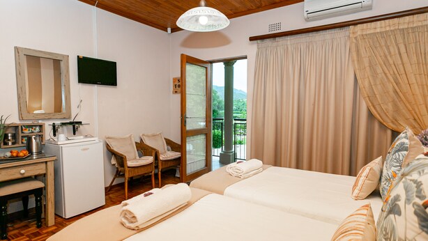 A hotel room with two beds, a desk with a refrigerator, a chair, and a view of greenery through the window. - Die Fonteine Country Guesthouse and Glamping (Oudtshoorn)